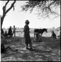 Man standing, with people sitting and standing near him, mule in the background, cattle in the distance