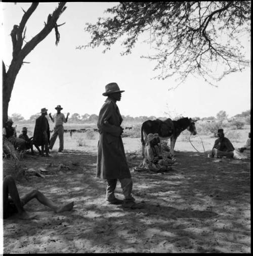 Man standing, with people sitting and standing near him, mule in the background, cattle in the distance