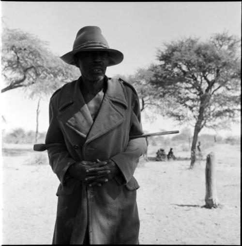 Man standing, with a group of people sitting near a tree in the background