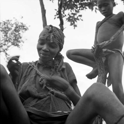 Woman wearing a kerchief and other ornaments on her head, working with a string, with a boy standing on one leg against a tree in the background