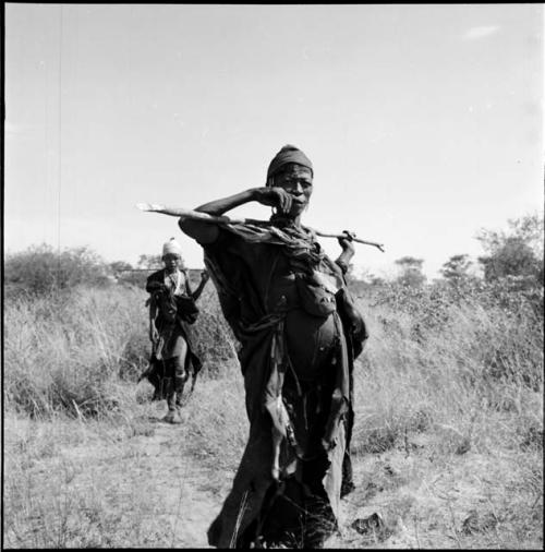 Woman standing, carrying a digging stick across her shoulders, with N!ai walking in the background