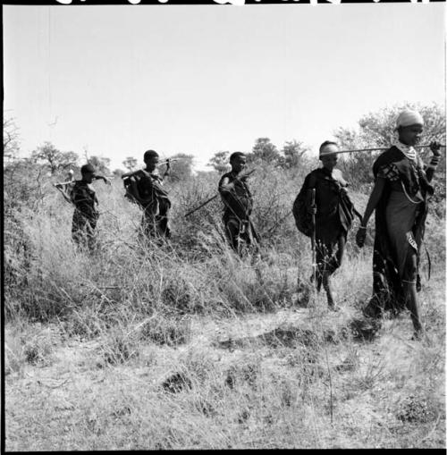 Five women walking in a line, carrying digging sticks on their shoulders