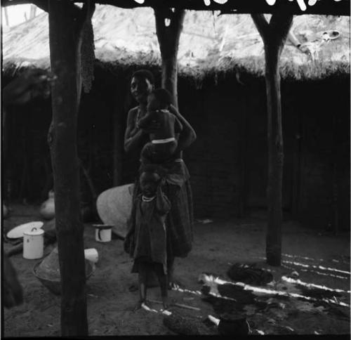 Woman holding a baby, standing with her child inside a skerm, with cooking equipment on the ground near them