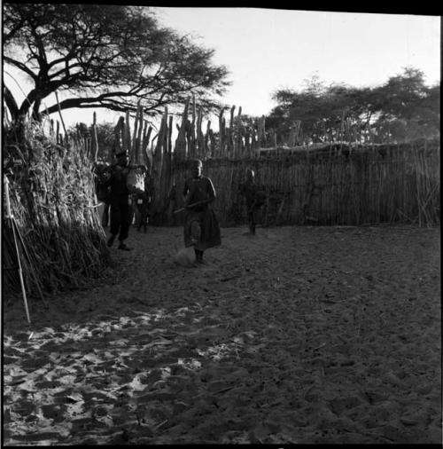 Man and a woman walking in front of a fence, with a woman holding a baby in the background