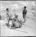 Di!ai squatting, fixing a girl's modesty apron, with another woman standing near them