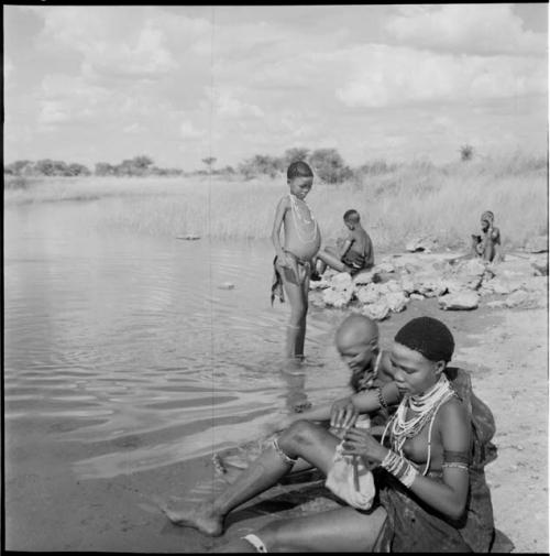 ≠Nisa sitting with a group of women at the edge of a pan, with a girl standing in the water next to them