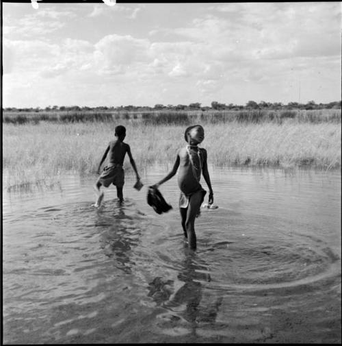 Girl standing in the water in a pan, with a boy wearing Western pants walking behind her, holding a tin can