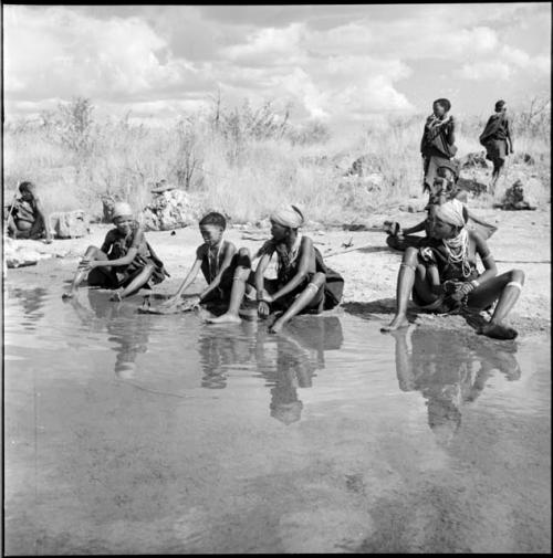 ≠Nisa sitting with other women at the edge of the pan, with a girl washing clothes, two women standing in the background