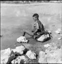 Woman wearing a kaross, sitting on a rock at the edge of a pan, bathing, with a digging stick on a rock next to her