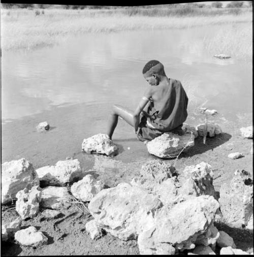 Woman wearing a kaross, sitting on a rock at the edge of a pan, bathing, with a digging stick on a rock next to her