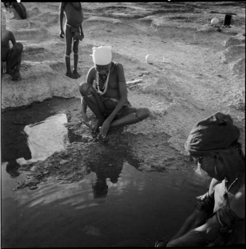 Woman sitting at the edge of a pan, washing her feet, with another woman sitting near her, child standing in the background, ostrich egg shells and a tin on the ground