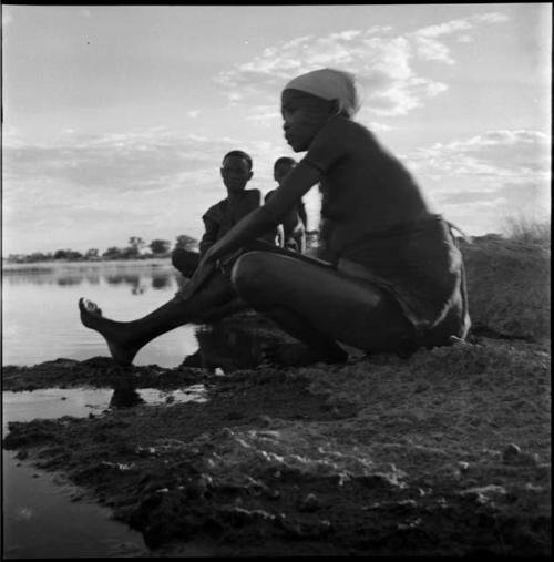 Woman sitting at the edge of a pan, washing her leg, with two people sitting in the background