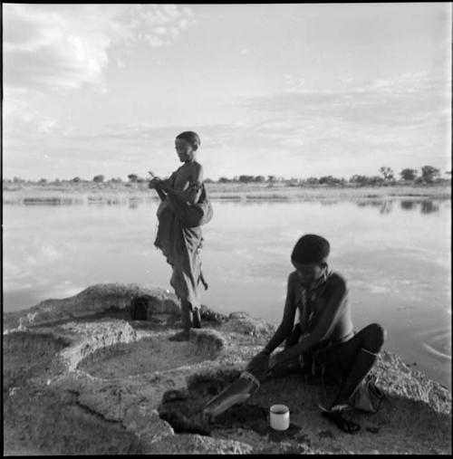 Woman sitting at the edge of a pan, washing her leg, with an enamel cup on the ground next to her, with another woman standing behind her