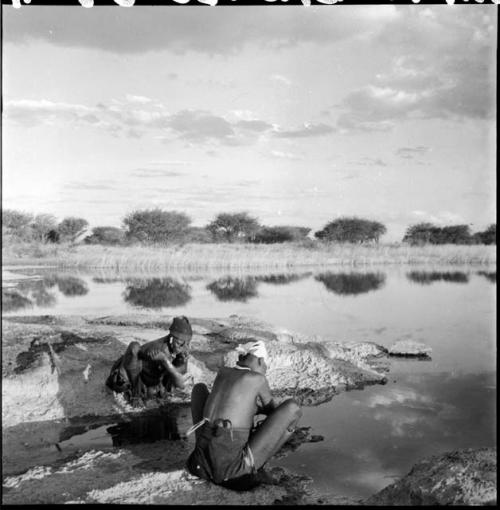 Di!ai and another woman squat at the edge of a pan, bathing