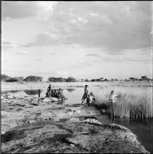 Group of people, including a man wearing Western clothing, sitting and standing at the edge of a pan