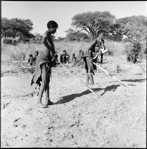 Two women beating mohengo, with a group of people sitting in the background