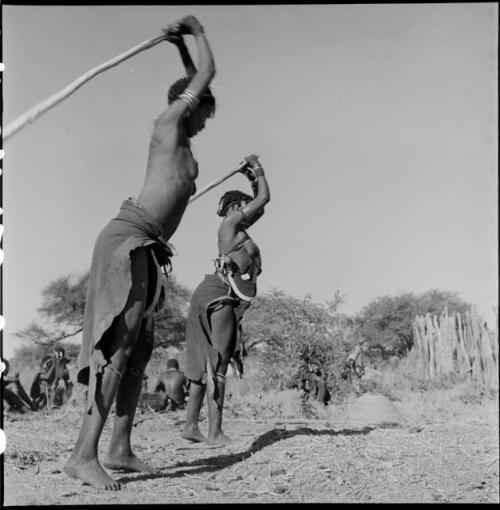 Two women beating mohengo with poles, with a group of people sitting in the background