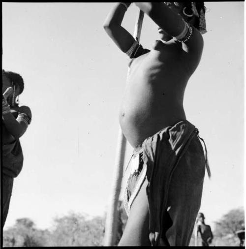 Woman standing, holding a pole used for beating mohengo, with another woman standing near her