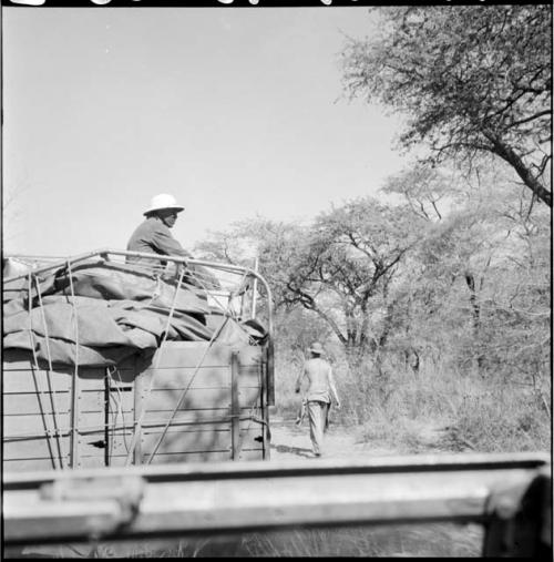 Man wearing a helmet, sitting in the back of an expedition truck view from the side, with another man walking away from the truck