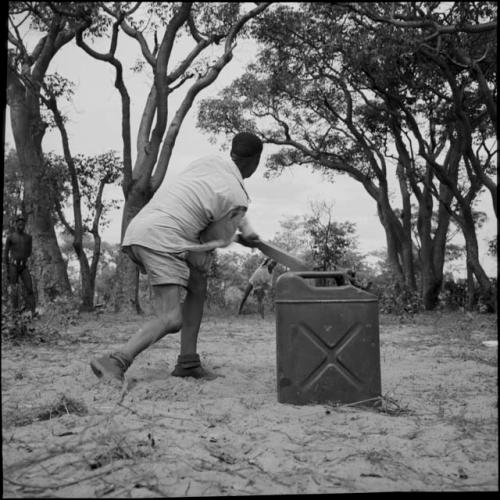 Man wearing Western clothing, swinging a bat in a baseball game, with a jerry can on the ground next to him, view from behind