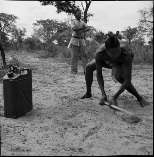 Man holding a bat in a baseball game, with a man wearing Western clothing standing behind him, jerry can on the ground next to him