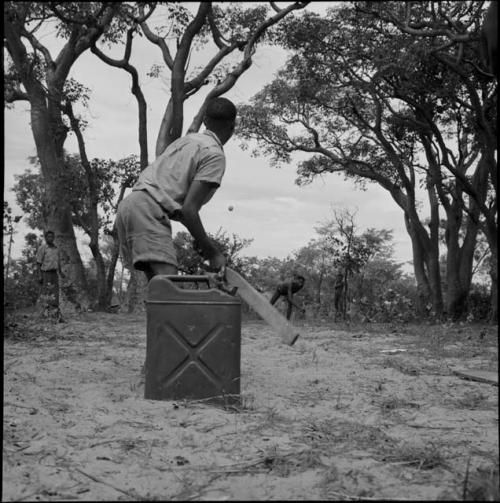 Man wearing Western clothing holding a bat in a baseball game, with a jerry can on the ground next to him