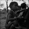 Boy sitting, with a woman with a child tied in her kaross sitting behind him