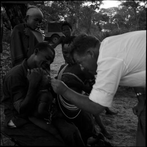 Child being fed medicine by Robert Story, sitting on Kernel Ledimo's lap, with N!ai and ≠Gisa watching