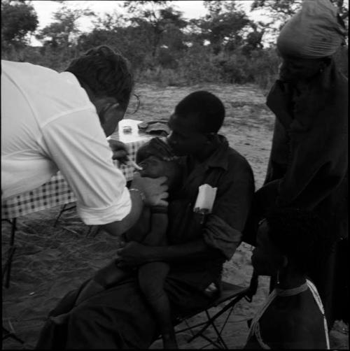 Baby being fed medicine by Robert Story, sitting on Kernel Ledimo's lap