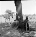 Two men sitting on the roots of a tree, with a horse standing next to them