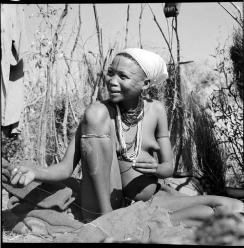 Woman sitting, stringing ostrich eggshell beads, with a string of beads on a piece of burlap next to her