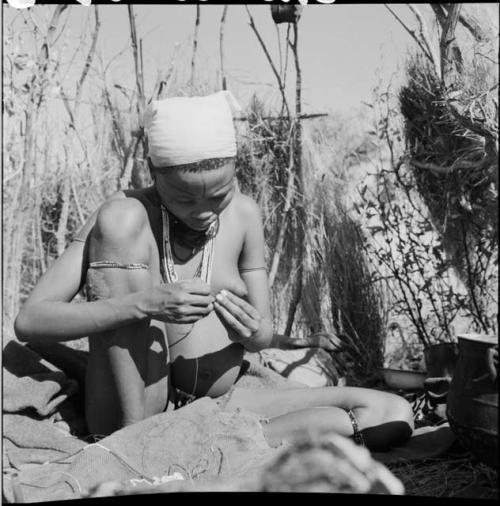 Woman sitting, stringing ostrich eggshell beads, with a string of beads on a piece of burlap next to her