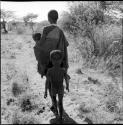 Woman carrying her baby tied on her back in her kaross, walking, with a child walking behind her, view from behind