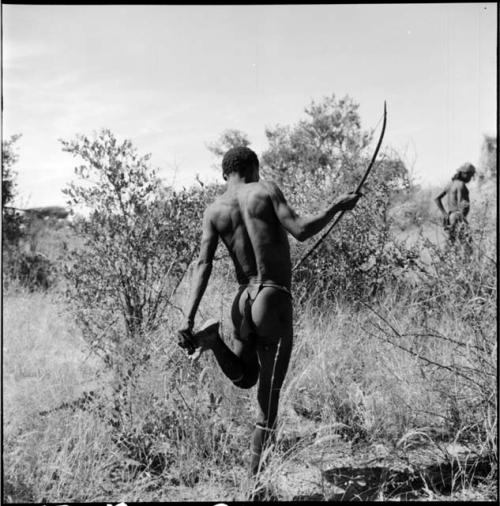 Bojo standing on one foot and taking a thorn out of his other foot, holding his bow