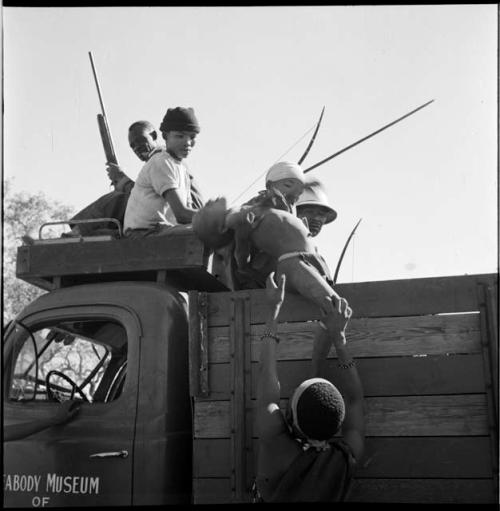 People sitting in the back of an expedition truck, some wearing Western clothing, holding bows, with a boy being lifted up into the truck