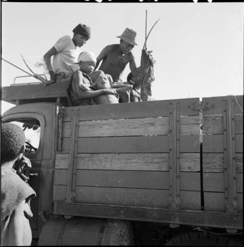 N!ai sitting in the back of an expedition truck, holding a boy, with men dressed in Western clothing and holding bows behind her