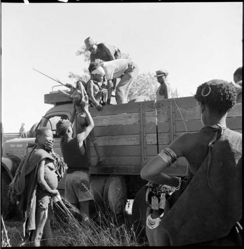 Man lifting a child down from the back of an expedition truck, with //Kushay and another woman standing near them, N!ai in the back of the truck with men wearing Western clothing