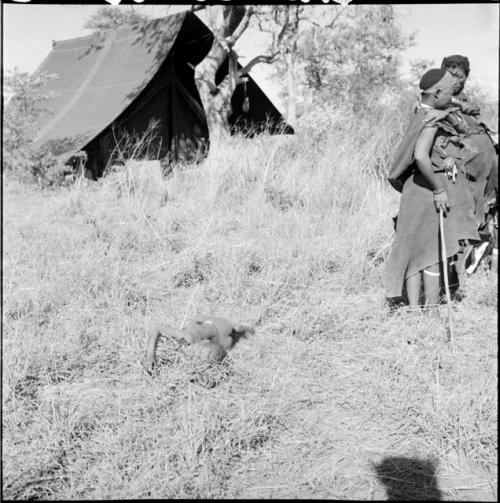 /Naoka and //Kushay standing with their backs to a boy sitting on the ground, crying, with an expedition tent in the background