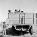 People standing or sitting in the back of an expedition truck, with a boy standing next to the truck, view from behind