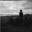 Man standing, holding a bow, with a group of men sitting behind him, pan in the distance