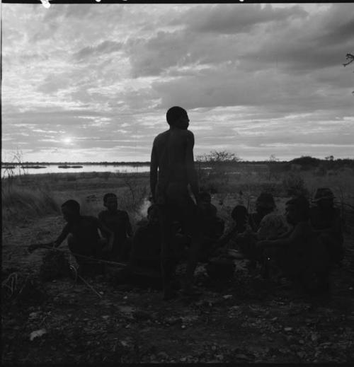 Man standing, holding a bow, with a group of men sitting behind him, pan in the distance