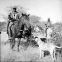 Two men riding horses, with a donkey and two dogs standing next to them