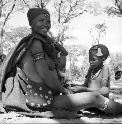 Woman wearing a kaross over one shoulder and a beaded bag over her other shoulder, sitting with a child in front of her