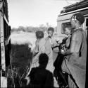 Younger woman helping /Naoka walk, with two other women standing next to them, between two expedition trucks; shadow of the photographer is visible