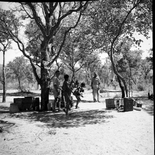 People standing with expedition members near the sound and film equipment, with enamel cups hanging from a line hung from the tree