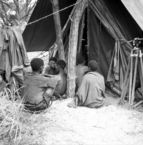 Group of people sitting next to an expedition tent, view from behind, with a camera on a tripod next to the tent