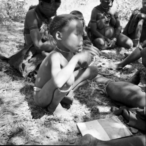 Boy squatting in front of a group of women sitting, with a magazine on the ground next to him
