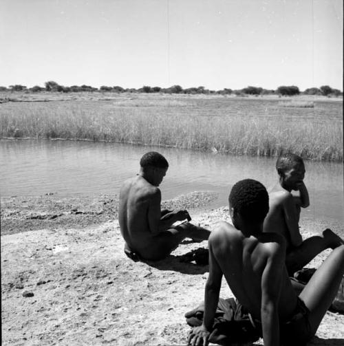 Three men sitting on the sand at the edge of a pan, view from behind