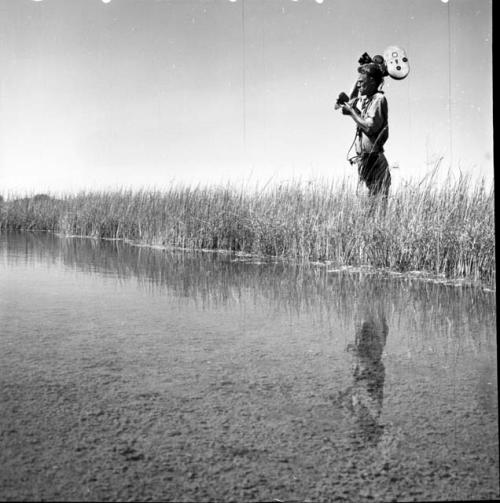 John Marshall standing in the grass at the edge of a pan, carrying his film camera on his shoulder