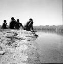 Man wrapped in a kaross, sitting with three other men on the sand at the edge of a pan